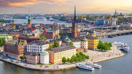 Aerial view of Stockholm&rsquo;s Gamla Stan at sunset, showing the historic island with colorful buildings, church spires, surrounding waterways, and boats docked along the quay.