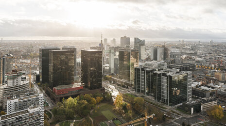 Aerial view of a modern city skyline with glass high-rise office towers clustered around green park areas, dense urban buildings stretching into the distance, and soft daylight breaking through cloudy skies.