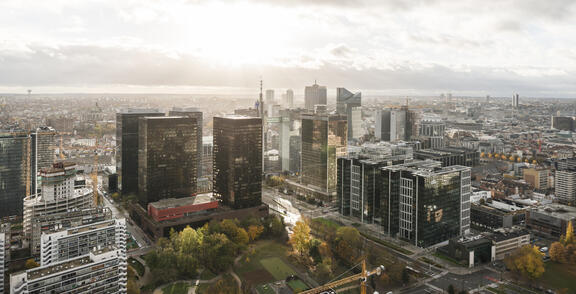 Aerial view of a modern city skyline with glass high-rise office towers clustered around green park areas, dense urban buildings stretching into the distance, and soft daylight breaking through cloudy skies.
