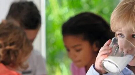 Child drinking a glass of milk while other children play and interact in the background.