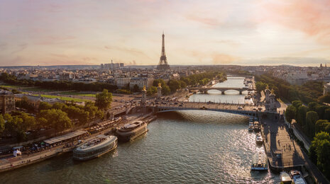 Aerial view of Paris at sunset with the Eiffel Tower in the distance, the River Seine flowing through the city, multiple historic bridges crossing the river, boats along the embankments, and warm golden light illuminating the urban landscape.