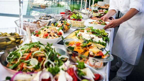 a buffet setup with a variety of fresh, colorful dishes, including salads, vegetables, and pickles. A person in a chef's uniform is arranging the food, ensuring everything is neatly displayed.
