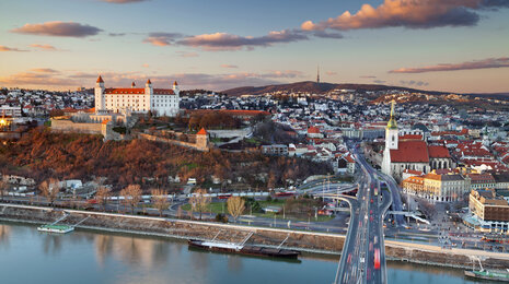 Aerial view of Bratislava at sunset, showing the Danube River, SNP Bridge, Bratislava Castle on the hill, and the historic old town with red-roofed buildings under a warm evening sky.