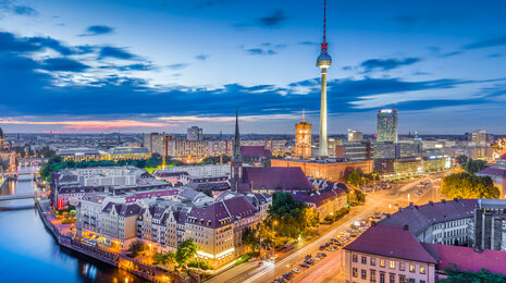 Evening cityscape of Berlin with the illuminated TV Tower (Fernsehturm) at Alexanderplatz, historic buildings along the Spree River, and city lights glowing at dusk under a blue sky.
