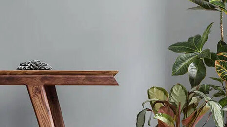 Minimalist interior scene with a wooden side table against a light gray wall, holding small decorative objects, next to a leafy indoor plant near a curtain.