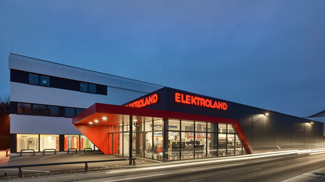 Elektroland electronics store exterior at night with illuminated signage and glass facade.