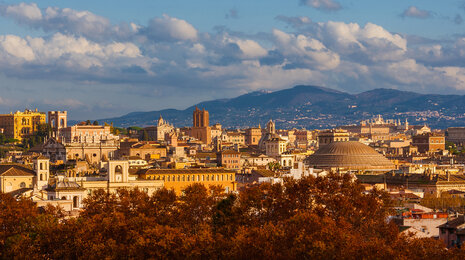 Panoramic view of Rome with historic buildings and domes spread across the city, warm sunlight illuminating the rooftops, autumn-colored trees in the foreground, and rolling hills under a partly cloudy sky in the background.
