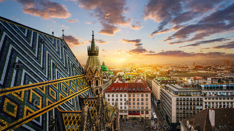 Sunset Vienna city view with ornate cathedral roof tiles in the foreground, historic spires, and a wide urban skyline glowing in warm evening light under dramatic clouds.