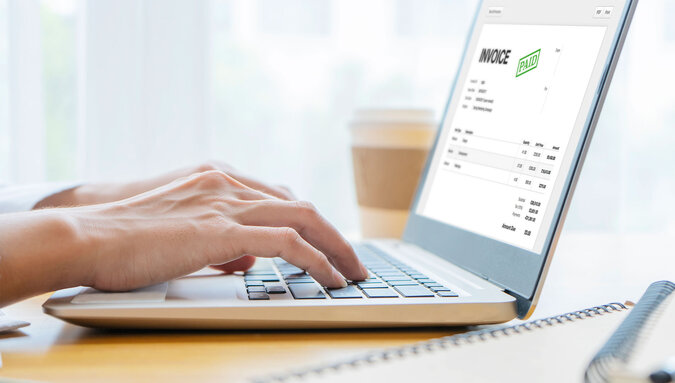 A person typing on a laptop displaying an invoice marked as 'PAID' on the screen
