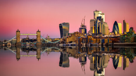 London skyline at sunset with Tower Bridge and modern skyscrapers reflected symmetrically in the calm River Thames under a pink and purple sky.