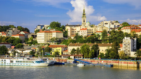 Riverside view of Belgrade&rsquo;s historic Zemun district, with colorful buildings on a green hillside, the Millennium Tower rising above the old town, and boats moored along the Danube under a clear blue sky.