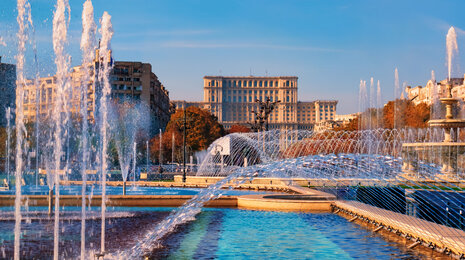 Large urban fountain with arcing water jets in the foreground, set in a city square with historic buildings and autumn-colored trees under a clear blue sky.
