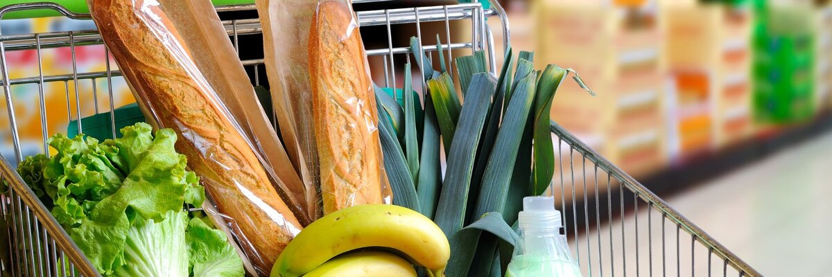 a shopping cart filled with groceries inside a supermarket. The cart contains fresh produce such as lettuce, tomatoes, leeks, bananas, and two baguettes. Additionally, there is a bottle of milk or juice, as well as other items like bread.
