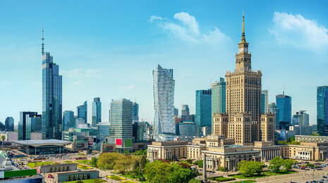Panoramic city skyline on a clear day, featuring the historic Palace of Culture and Science at center, surrounded by modern skyscrapers, green parks, and busy urban streets.