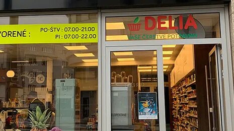 Entrance of a DELIA grocery store with glass doors, visible shelves inside, and opening hours displayed above the entrance.