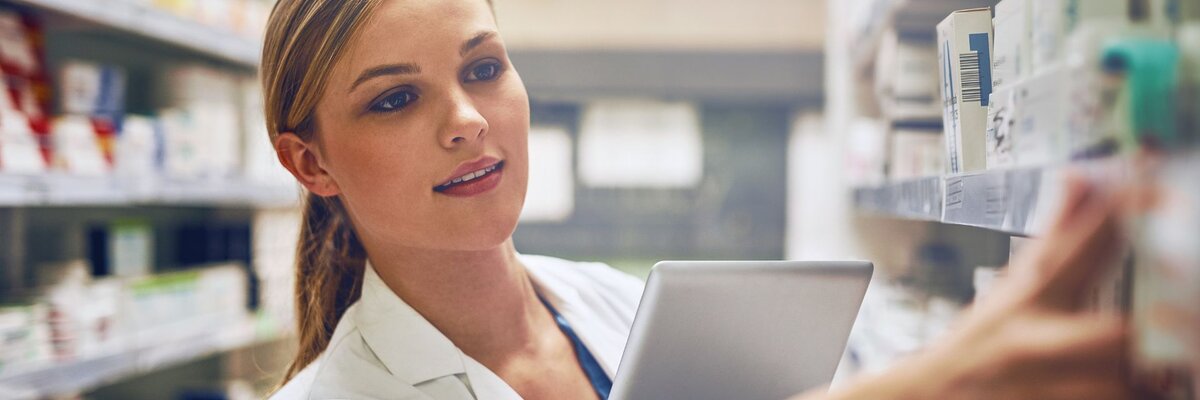 Female pharmacist organizing medications on a shelf while holding a tablet for inventory management in a pharmacy