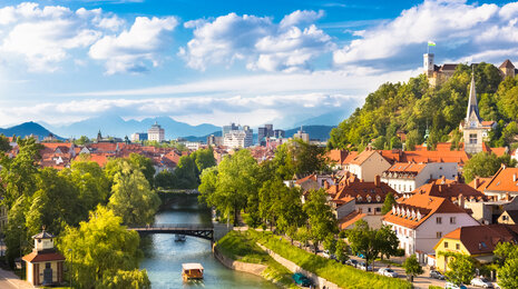 Sunny panoramic view of Ljubljana, Slovenia, with the Ljubljanica River winding through the historic old town, red-roofed buildings, green trees, and Ljubljana Castle on the hill under a blue sky.
