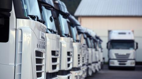 Close-up of a row of white trucks parked in a logistics yard, with one truck in focus in the background, emphasizing fleet management and transportation