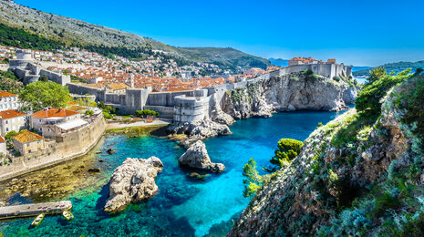 Sunlit coastal city with medieval stone walls perched above crystal-clear turquoise water, rocky cliffs in the foreground, and red-roofed historic buildings stretching along the shoreline under a bright blue sky.