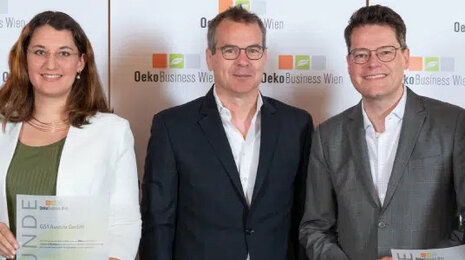 Group of five professionally dressed business representatives standing in front of a branded backdrop at an award or recognition event, smiling and holding certificates.