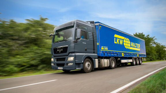 Blue LKW WALTER semi-truck driving on a rural road, captured in motion with blurred greenery in the background, representing long-distance logistics and road transport.