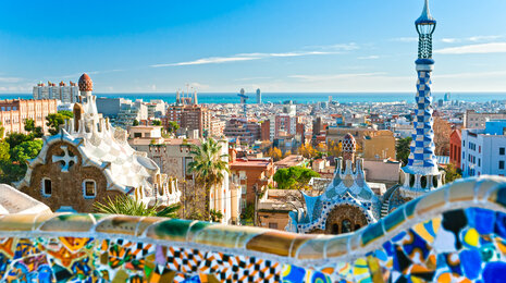 Panoramic view of Barcelona from Park G&uuml;ell, featuring colorful mosaic terraces in the foreground, Gaud&iacute;-style towers and rooftops, and the city skyline stretching toward the Mediterranean Sea under a clear blue sky.