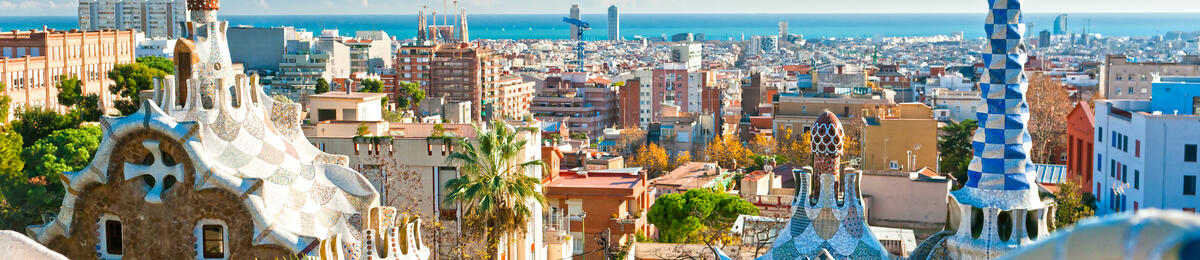 Panoramic view of Barcelona from Park Güell, featuring colorful mosaic terraces in the foreground, Gaudí-style towers and rooftops, and the city skyline stretching toward the Mediterranean Sea under a clear blue sky.