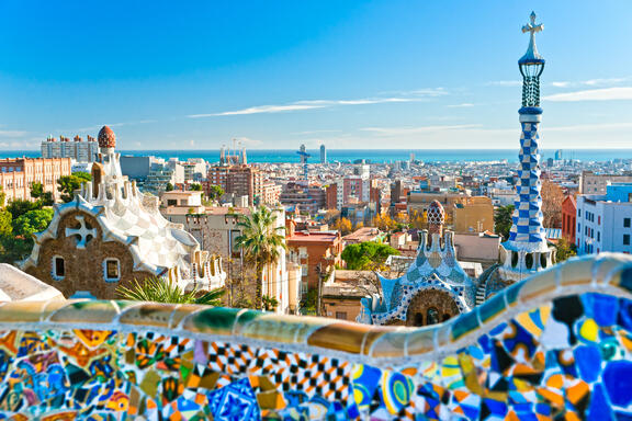 Panoramic view of Barcelona from Park Güell, featuring colorful mosaic terraces in the foreground, Gaudí-style towers and rooftops, and the city skyline stretching toward the Mediterranean Sea under a clear blue sky.