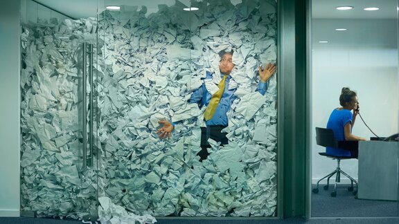 A man trapped in an office overflowing with crumpled paper, symbolizing being overwhelmed by paperwork, while a woman in the adjacent room speaks calmly on the phone