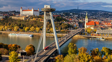 Aerial view of the SNP Bridge (UFO Bridge) spanning the Danube River in Bratislava, with the UFO observation deck, Bratislava Castle on the hill, and autumn trees along the riverbanks.