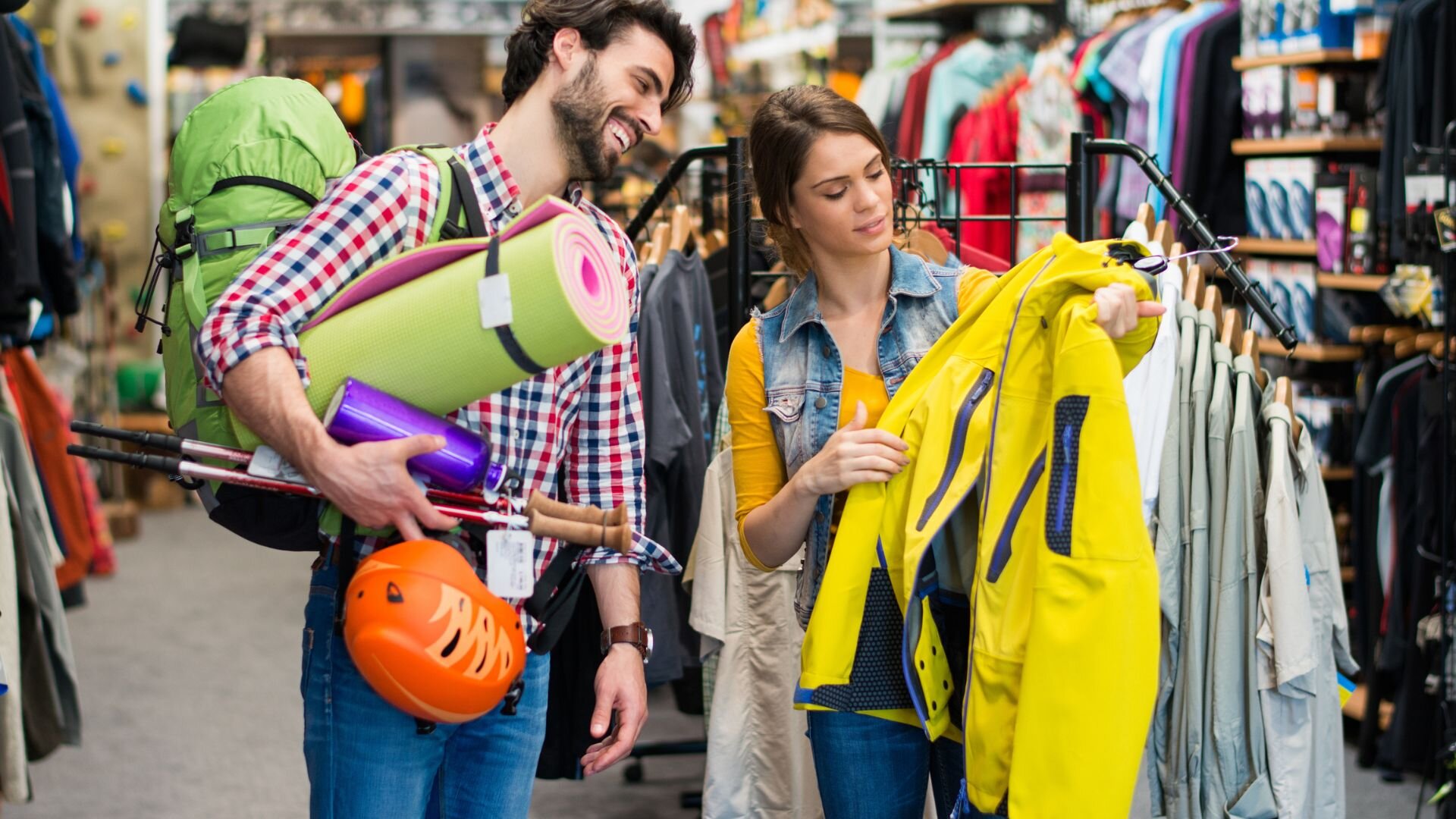a couple shopping for outdoor gear in a retail store. The man is smiling and carrying various camping and hiking equipment, including a large green backpack, trekking poles, a helmet, and a sleeping mat. The woman is holding up a yellow jacket, inspecting it closely