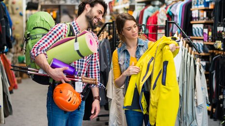 a couple shopping for outdoor gear in a retail store. The man is smiling and carrying various camping and hiking equipment, including a large green backpack, trekking poles, a helmet, and a sleeping mat. The woman is holding up a yellow jacket, inspecting it closely