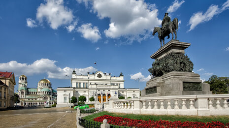 Equestrian statue on a stone pedestal in a grand city square, with a historic opera house and domed cathedral in the background under a blue sky with scattered clouds.